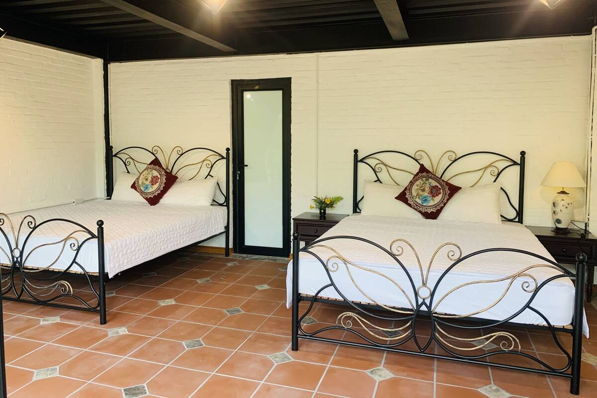 Una habitación con dos camas, cada una con estructura de metal negro y ropa de cama blanca. Hay una mesita con una lámpara y un florero. Las paredes son blancas y el suelo está cubierto de baldosas.