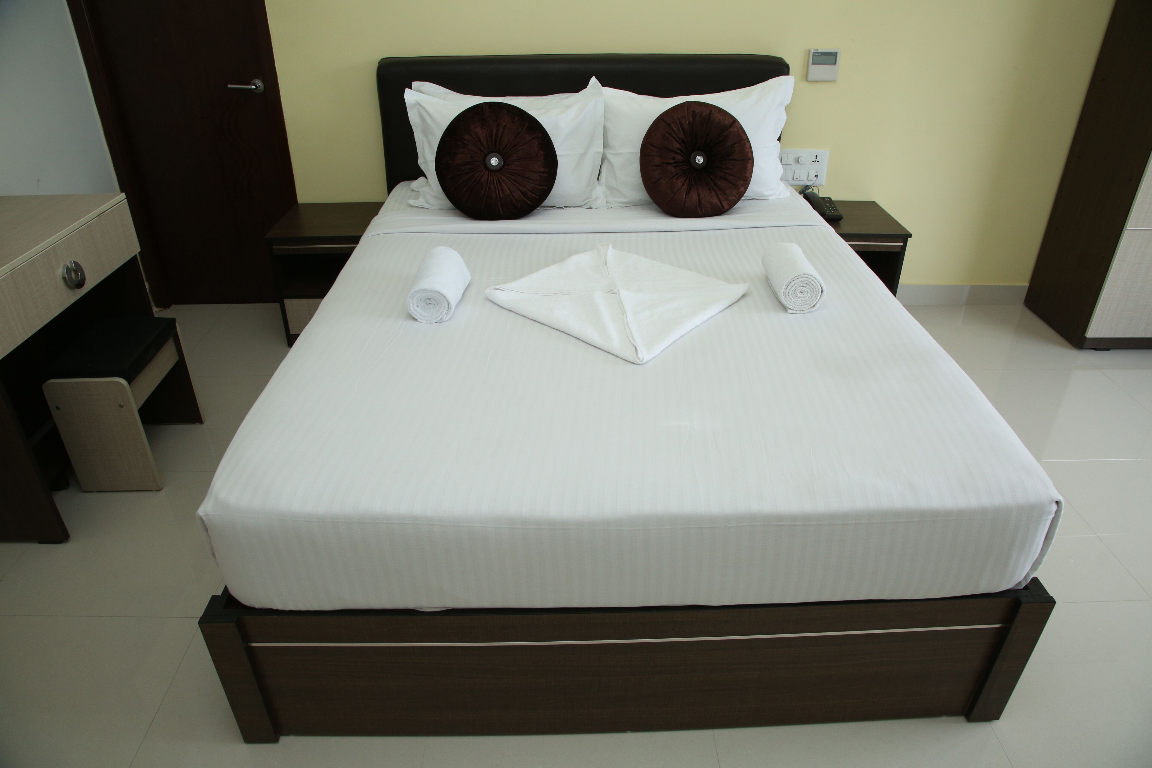 Una cama bien hecha con sábanas blancas, dos almohadones decorativos y toallas enrolladas sobre un cabecero de madera oscura en una habitación de hotel.