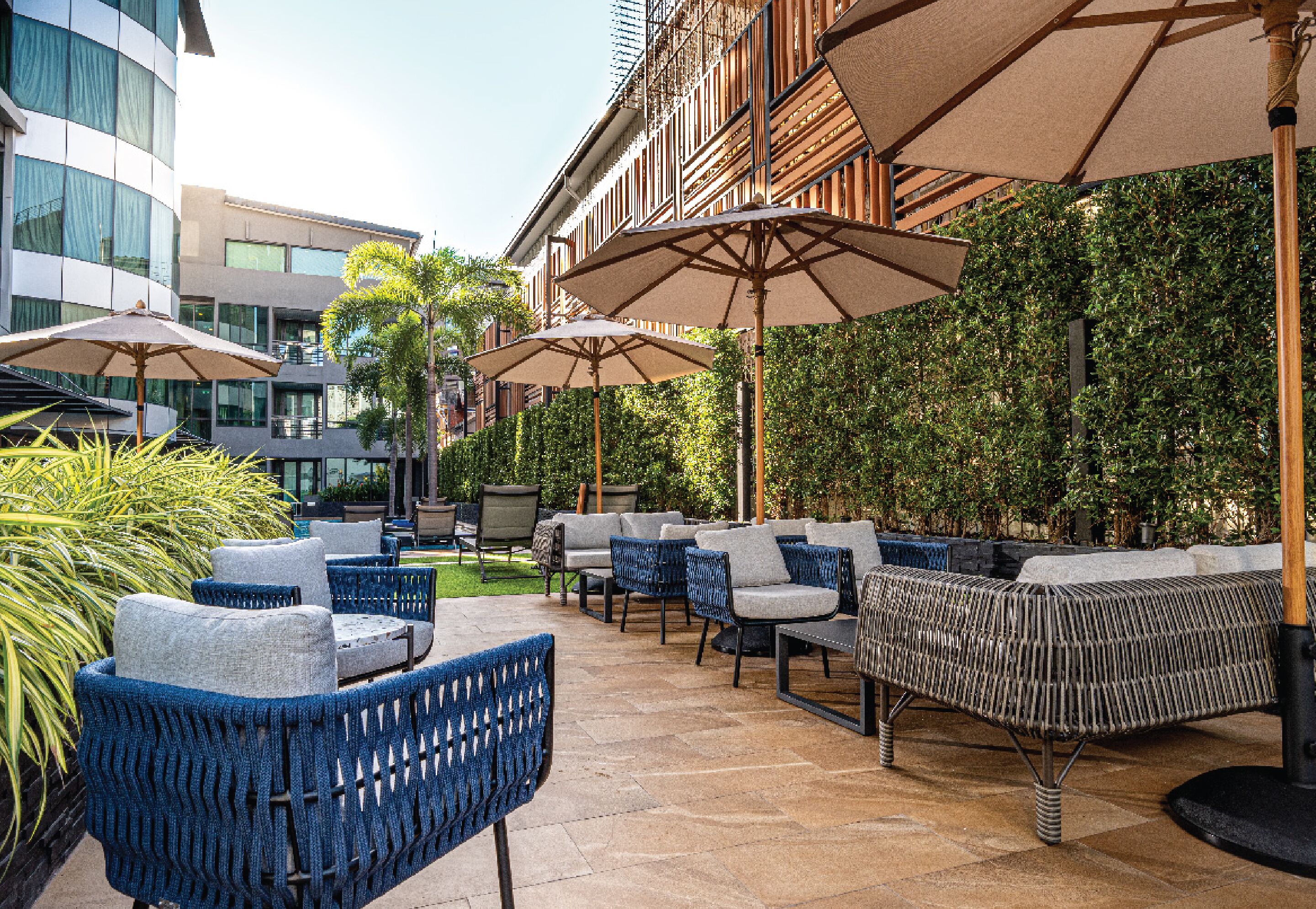 Un espace extérieur aménagé avec des meubles en osier et des parasols, entouré de verdure et de bâtiments modernes.