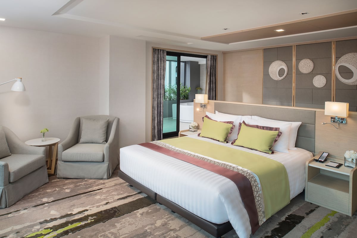 A modern hotel room with a large bed, a sitting area with a chair and a small table, a nightstand with a lamp, and a view of the outdoors through a sliding door.