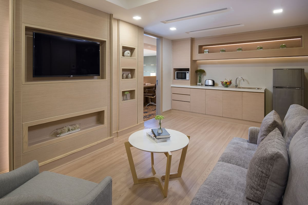 A modern living room with a flat-screen TV, a grey sofa, a small white table with a book and vase, and a kitchen area with a microwave and refrigerator.