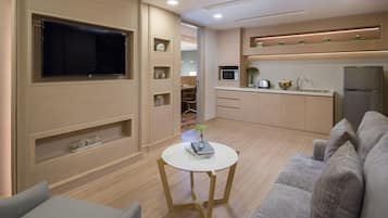 Una sala de estar moderna con un televisor de pantalla plana, un sofá gris, una mesita blanca con un libro y un jarrón, y una zona de cocina con microondas y refrigerador.