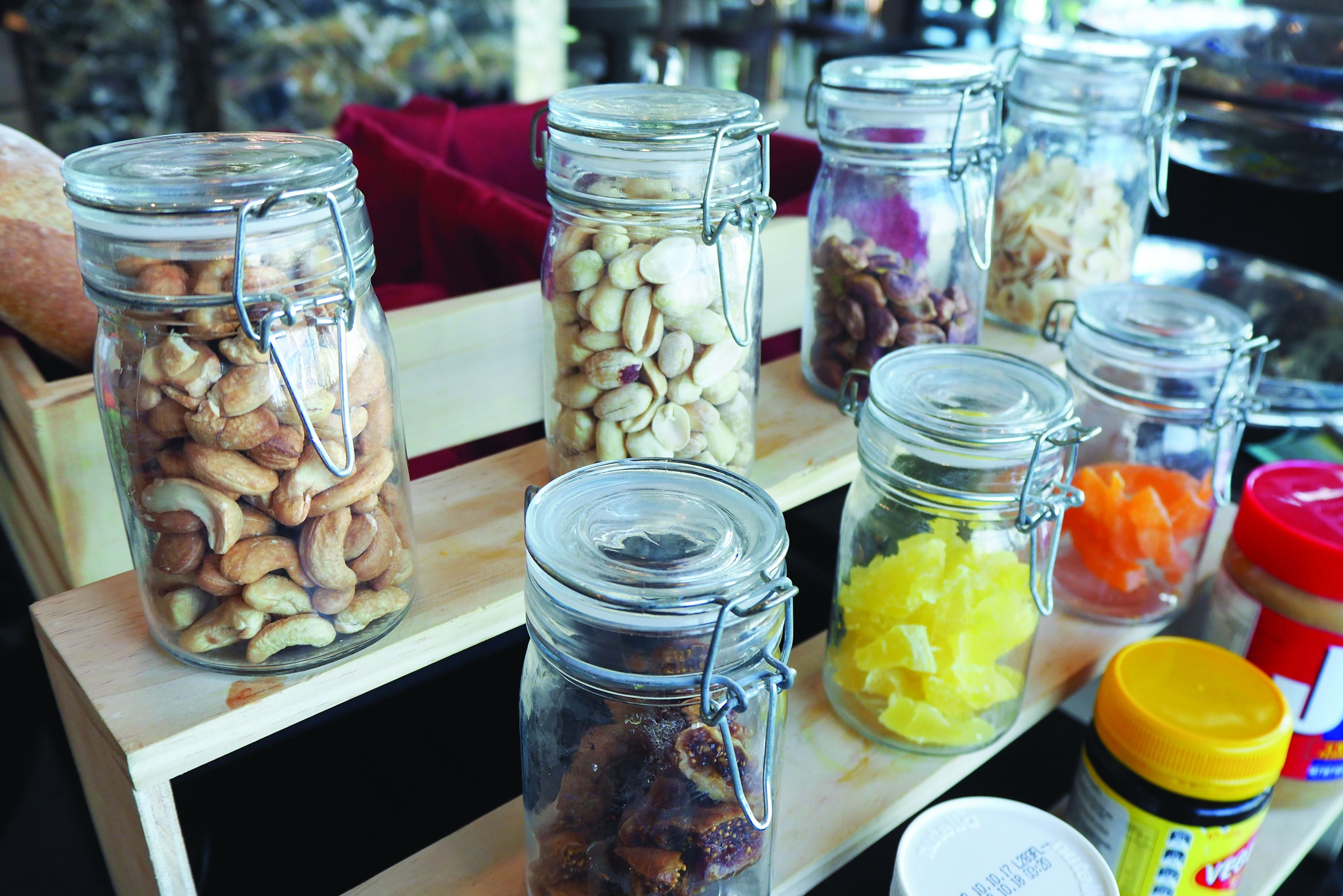 On peut voir une variété de bocaux en verre contenant différents aliments, comme des noix, des fruits secs et des légumes, disposés sur une étagère en bois.