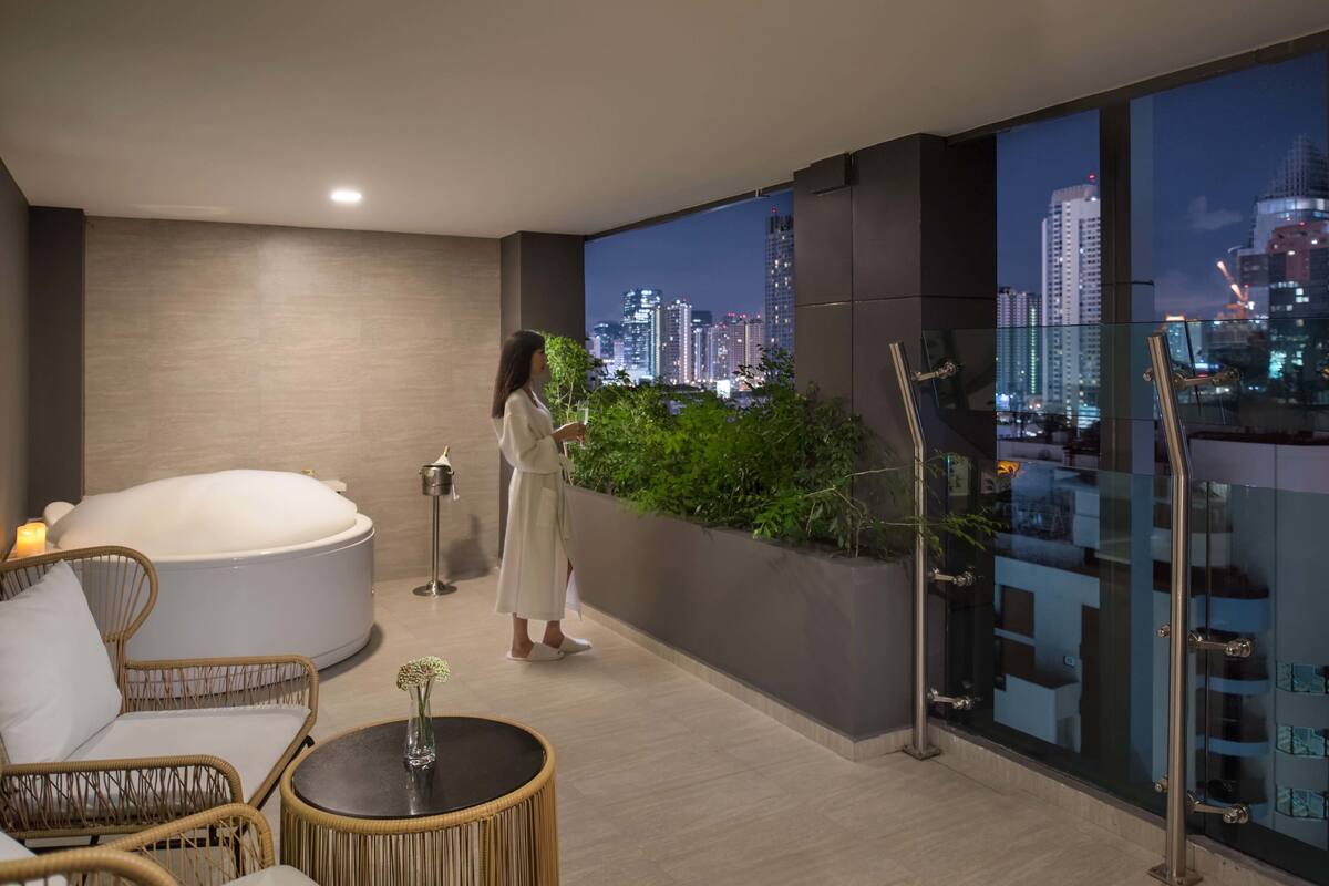 A modern room with a large bathtub, a glass wall with a city view, and a small table with a vase.
