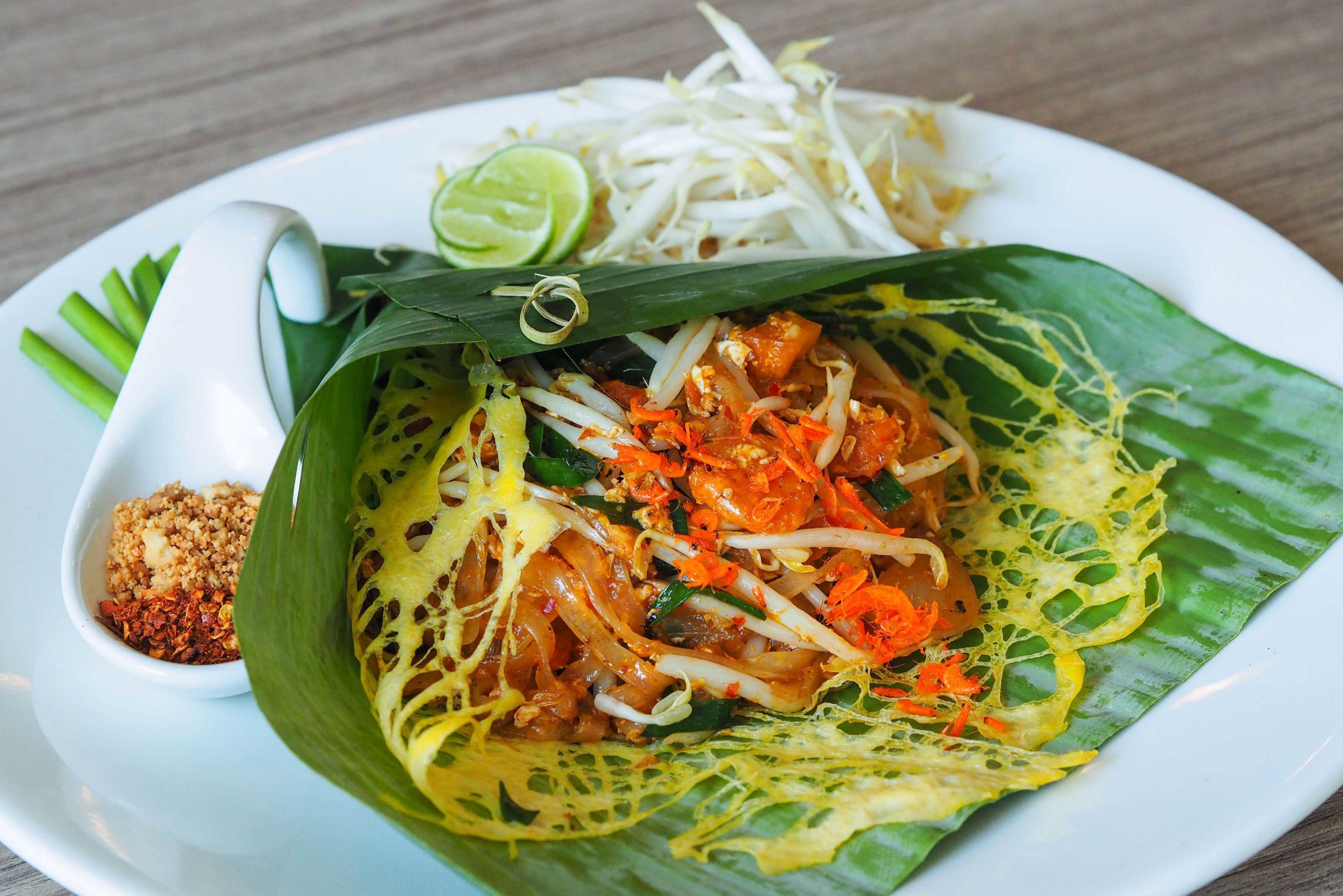 A plate of Pad Thai with bean sprouts and lime, served on a white plate with a small bowl of chili and peanuts.