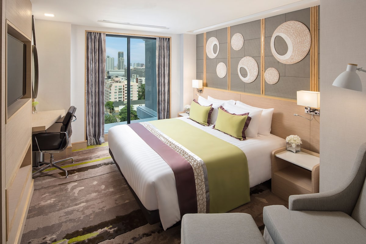 A hotel room with a large bed, a desk, a chair, a TV, and a view of the city.