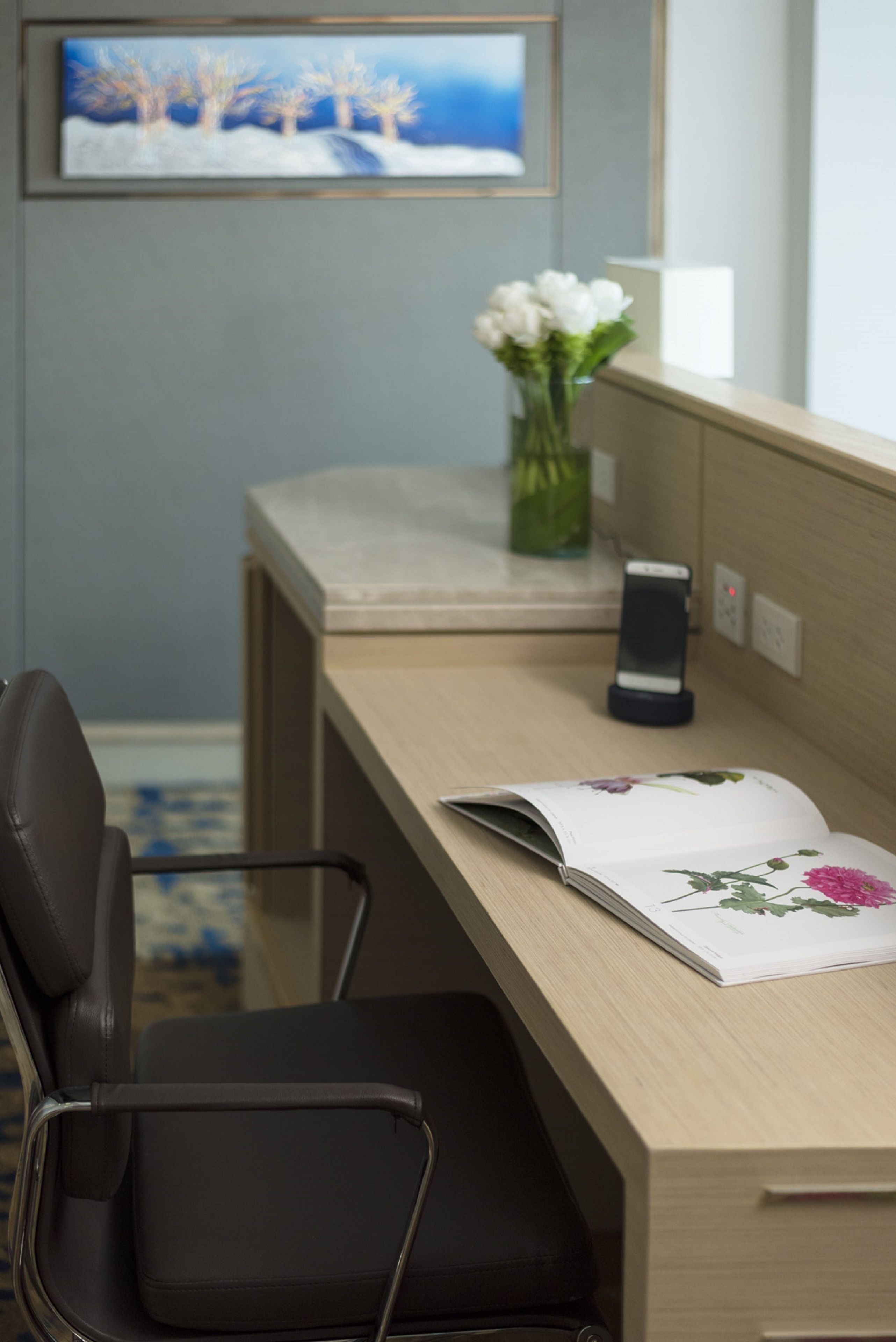 Ein Hotelzimmer mit einem Holztisch, einem schwarzen Bürostuhl, einer Vase mit weißen Blumen, einem gerahmten Gemälde an der Wand und einem Telefon auf dem Tisch.