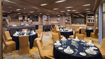 Comedor de hotel con mesas redondas dispuestas para una comida, sillas y estanterías con libros y objetos decorativos.