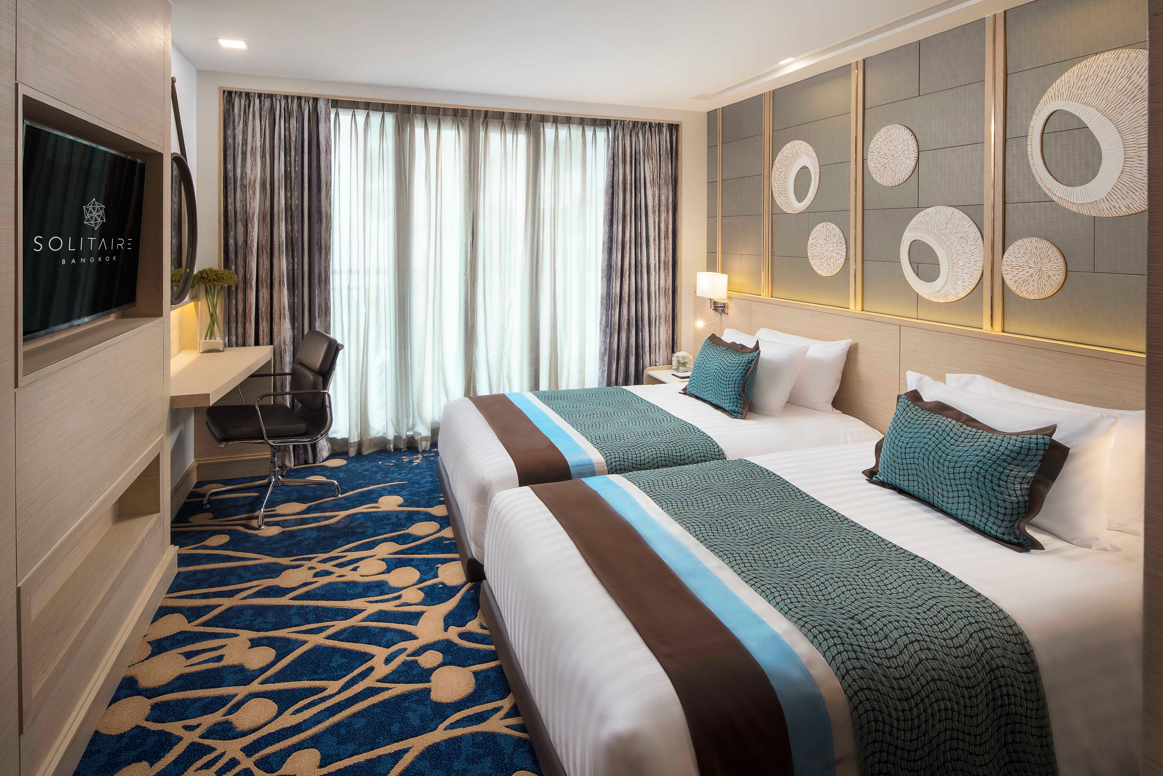 A hotel room with two beds, a desk, a chair, a TV, and a large window with curtains.