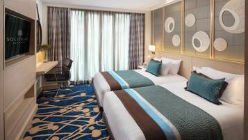 A hotel room with two beds, a desk, a chair, a TV, and a large window with curtains.