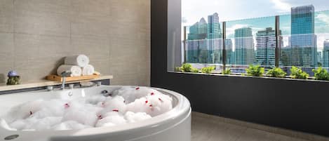 A modern bathroom with a large bathtub filled with bubbles, a window with a city view, and a shelf with towels and toiletries.