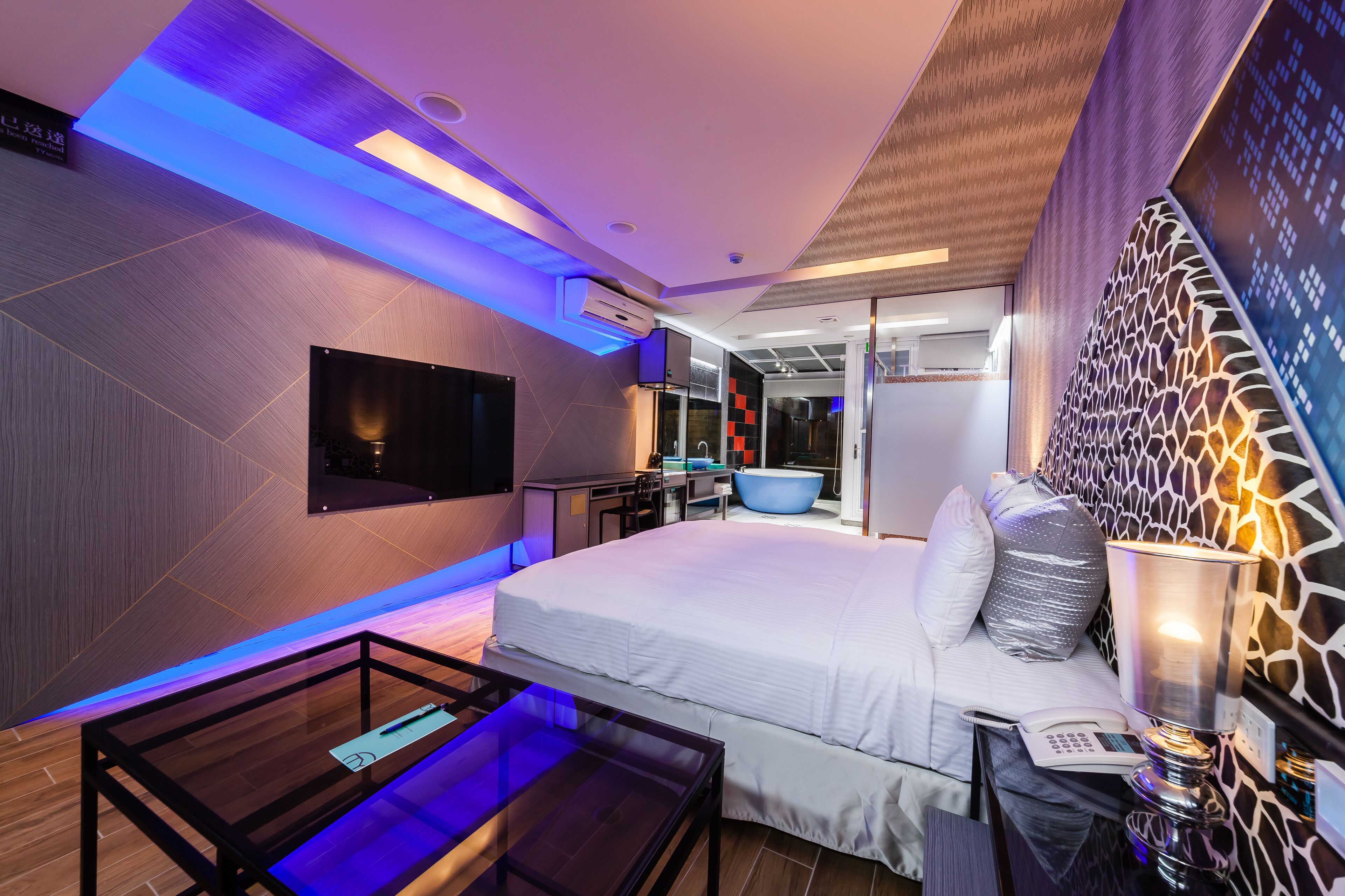 A modern hotel room with a large bed, a glass coffee table, a flat-screen TV, a bathroom with a bathtub, and a unique wall design.