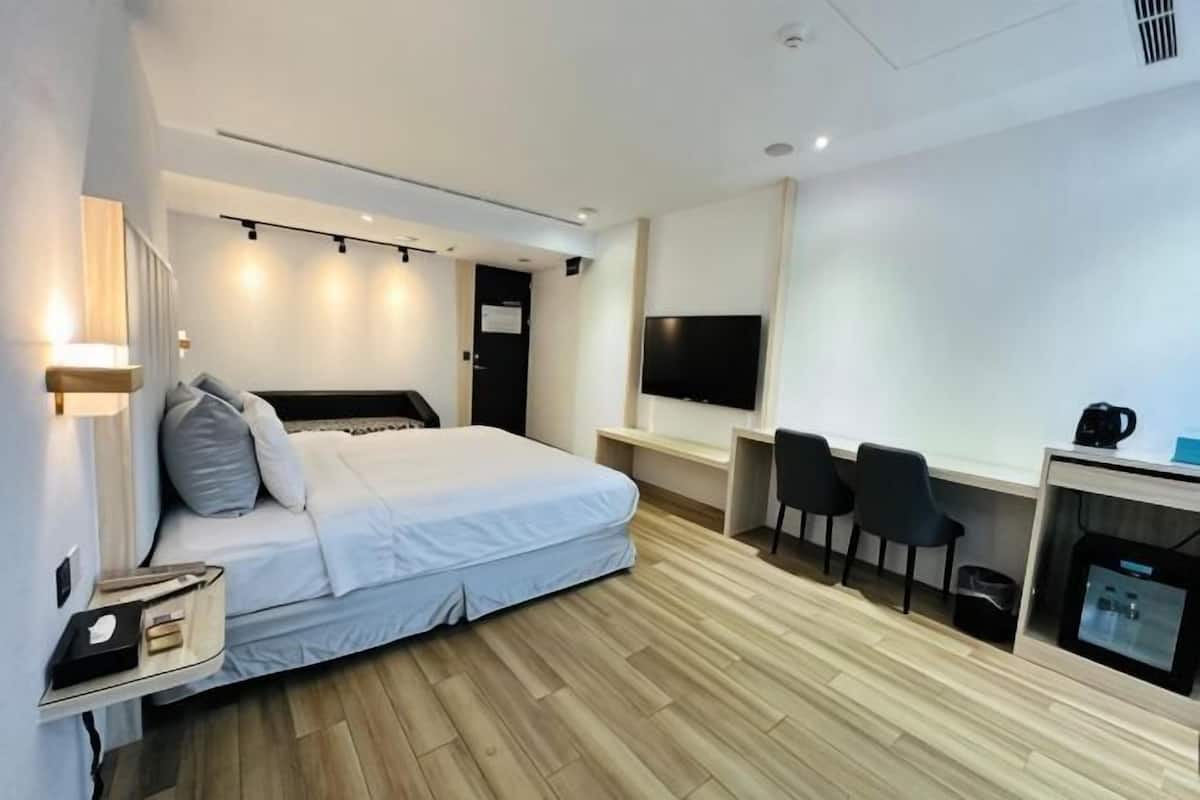 A modern hotel room with a bed, bedside table, TV, desk, chairs, and a coffee machine.