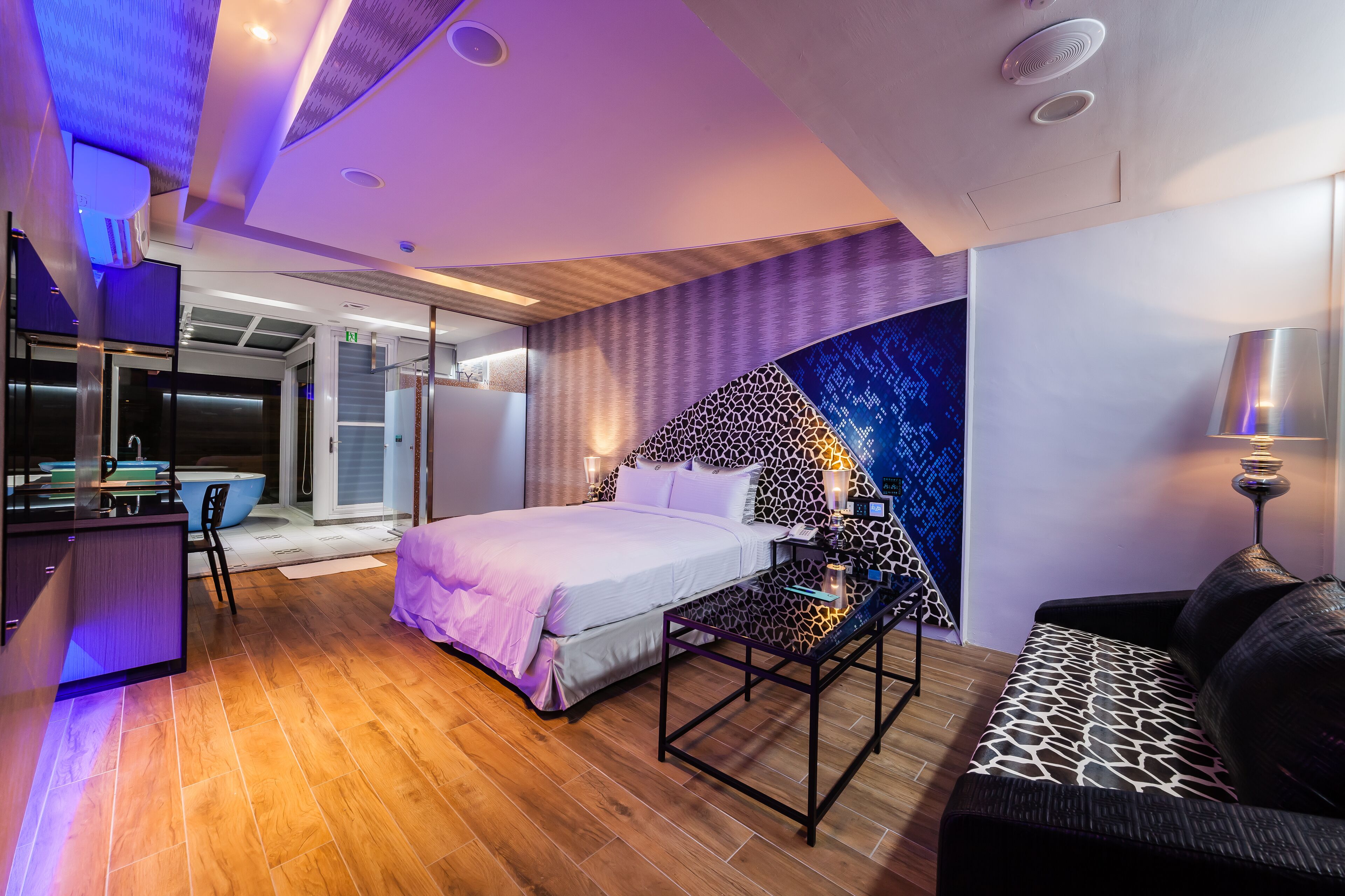 A modern hotel room with a bed, a desk, and a sofa. There is a bathroom with a glass door and a shower.