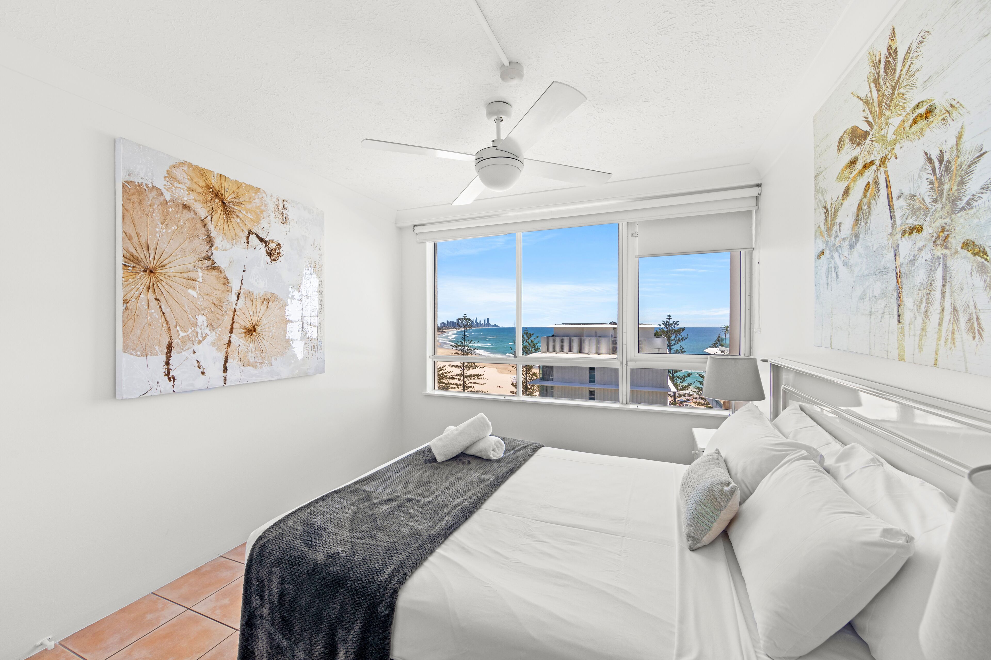 A bedroom with a bed, a ceiling fan, a window with a view of the beach, and two wall art pieces.