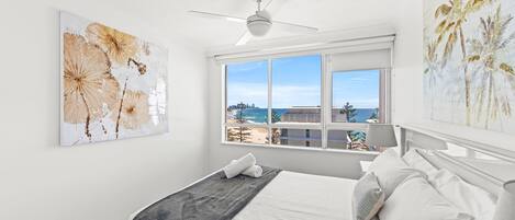 A bedroom with a bed, a ceiling fan, a window with a view of the beach, and two wall art pieces.