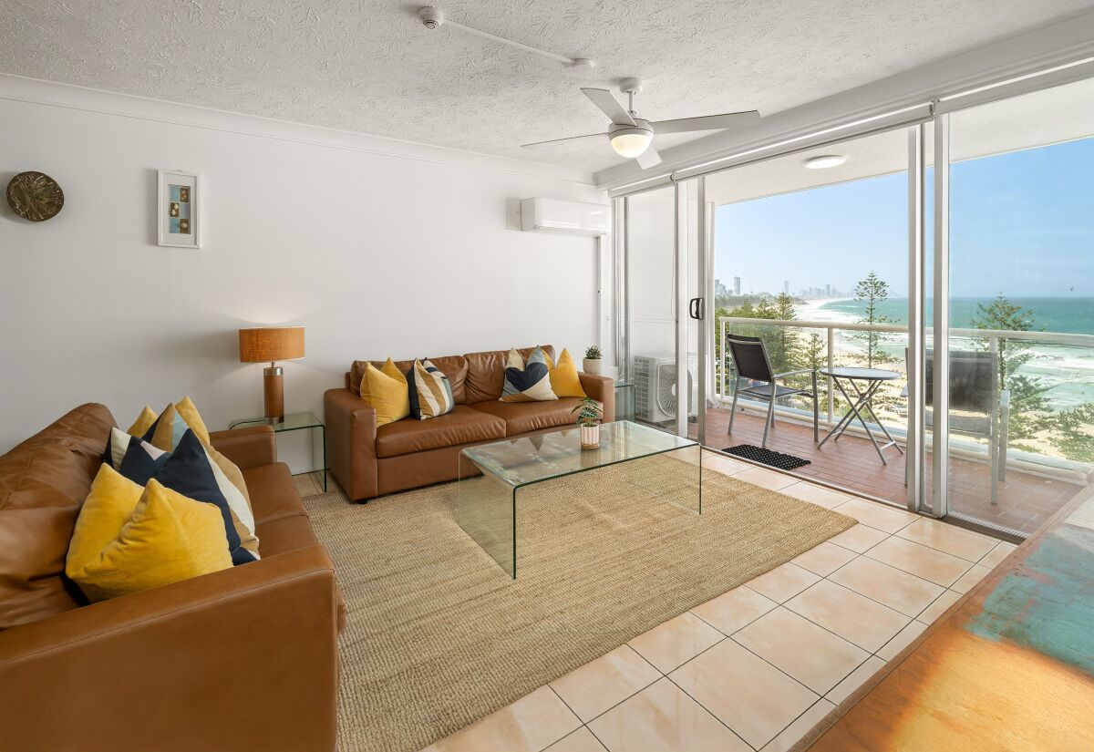 A living room with a brown sofa, yellow pillows, a glass coffee table, and a view of the beach through large sliding glass doors.