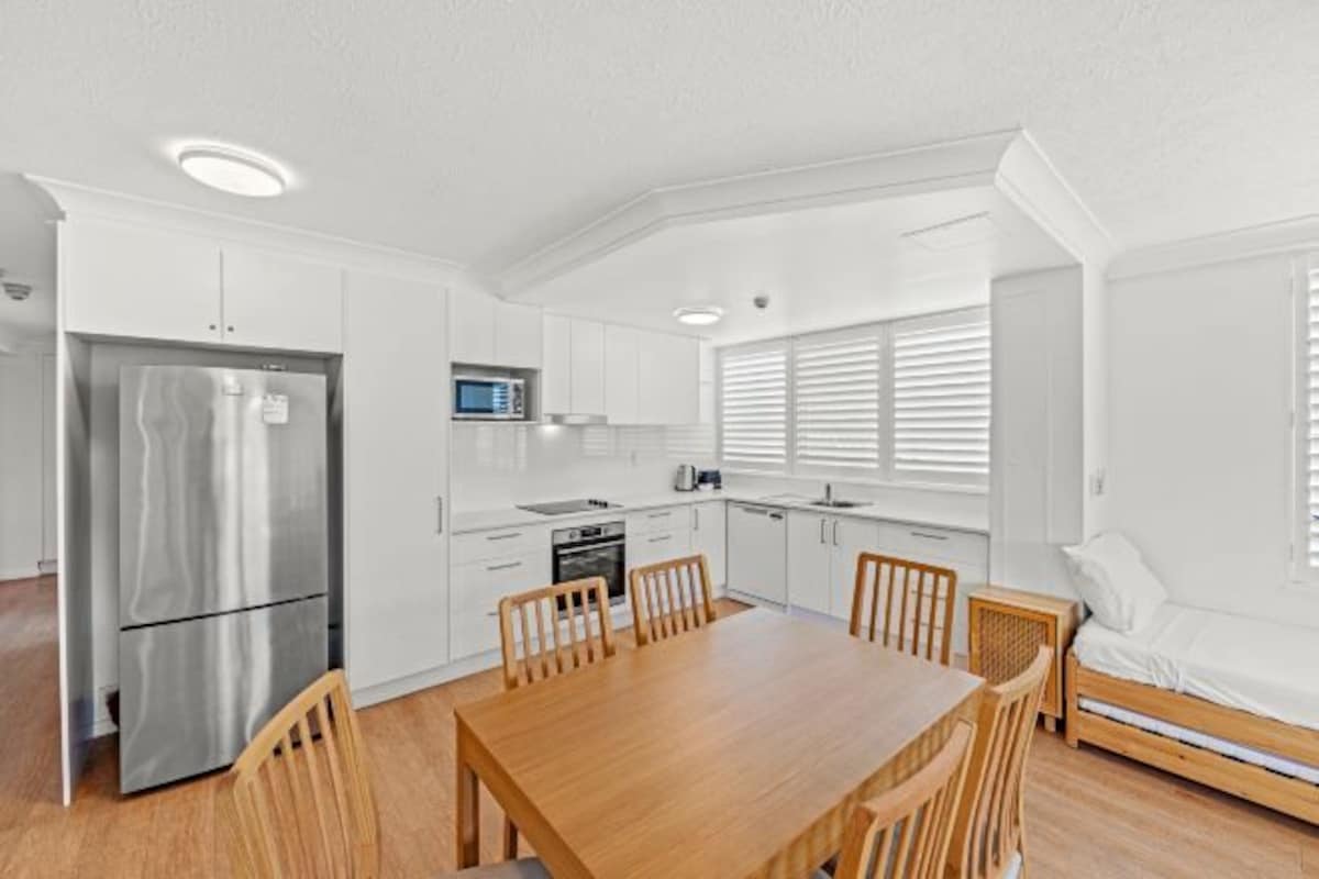 A compact kitchen with white cabinets, a stainless steel refrigerator, a microwave, and a dining table with chairs.
