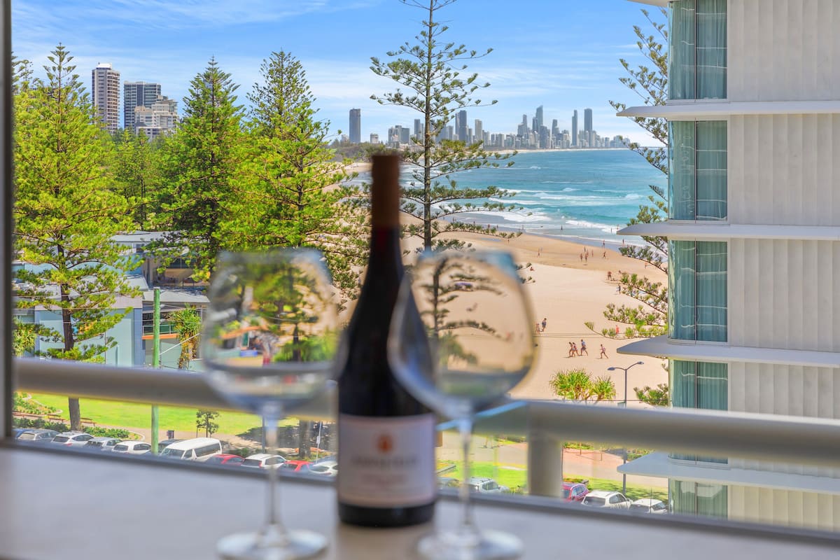 A bottle of wine and two wine glasses on a balcony overlooking a beach with a city skyline in the background.