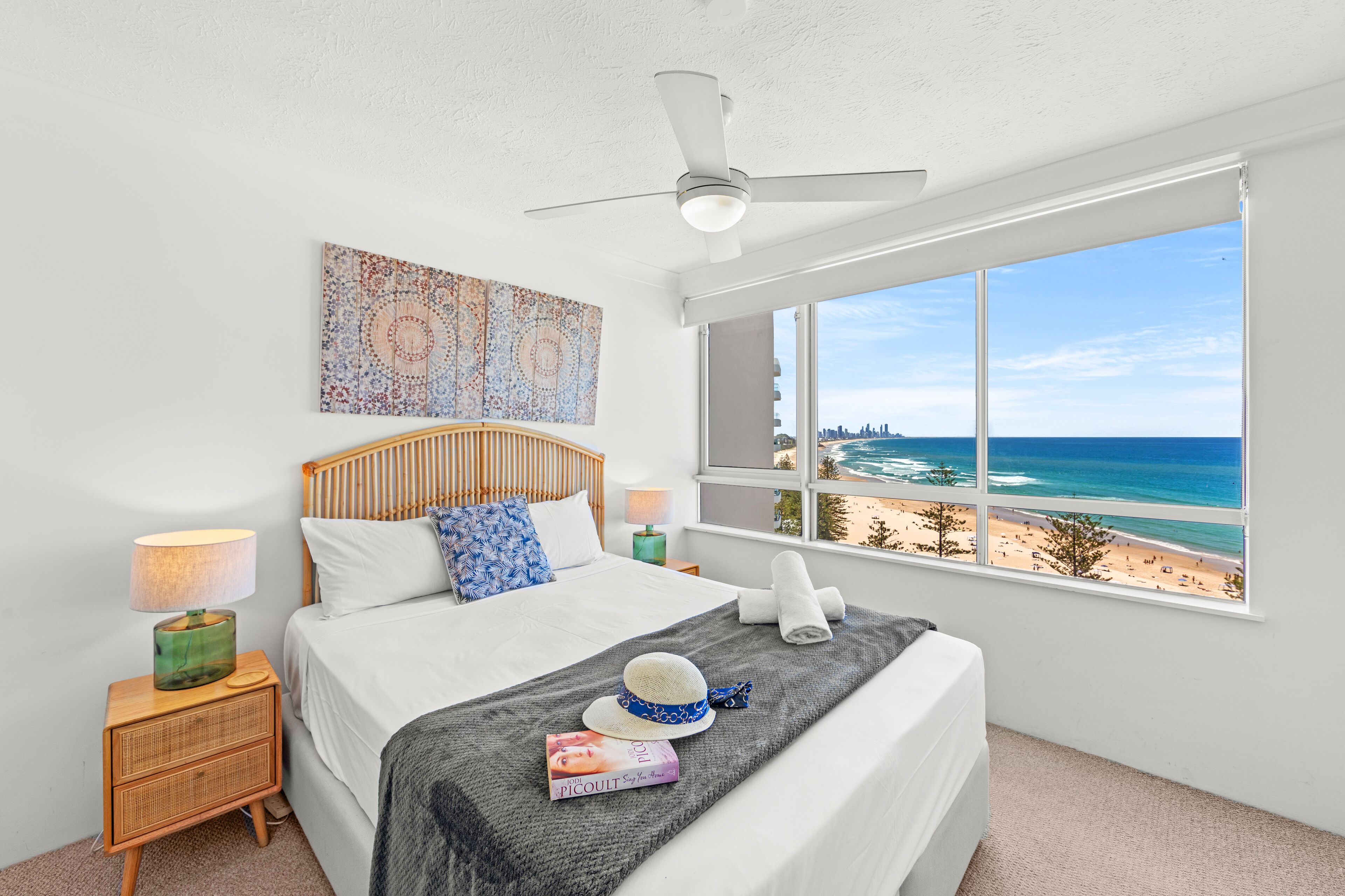 Un dormitorio con cama, mesita de noche, lámpara, ventana con vista a la playa y ventilador de techo.