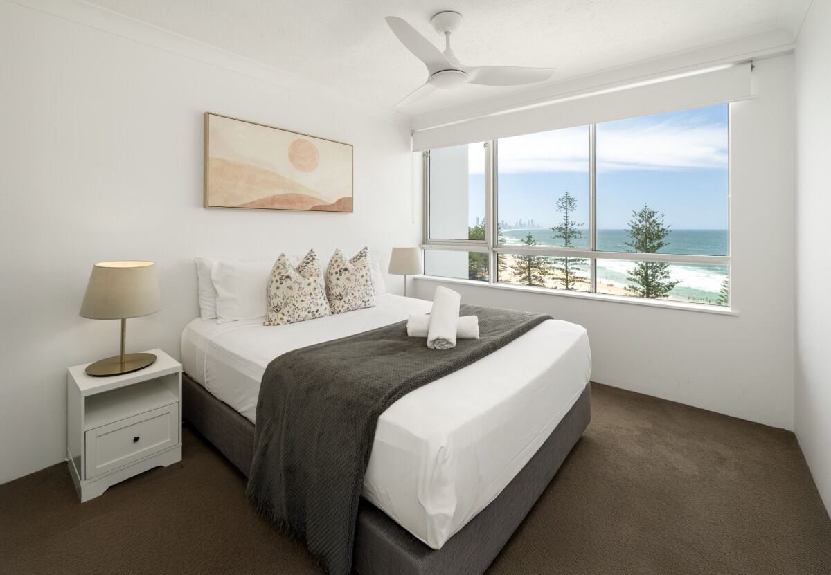 Een slaapkamer met een bed, bedlampen, een plafondventilator, een raam met uitzicht op het strand, en een schilderij aan de muur.