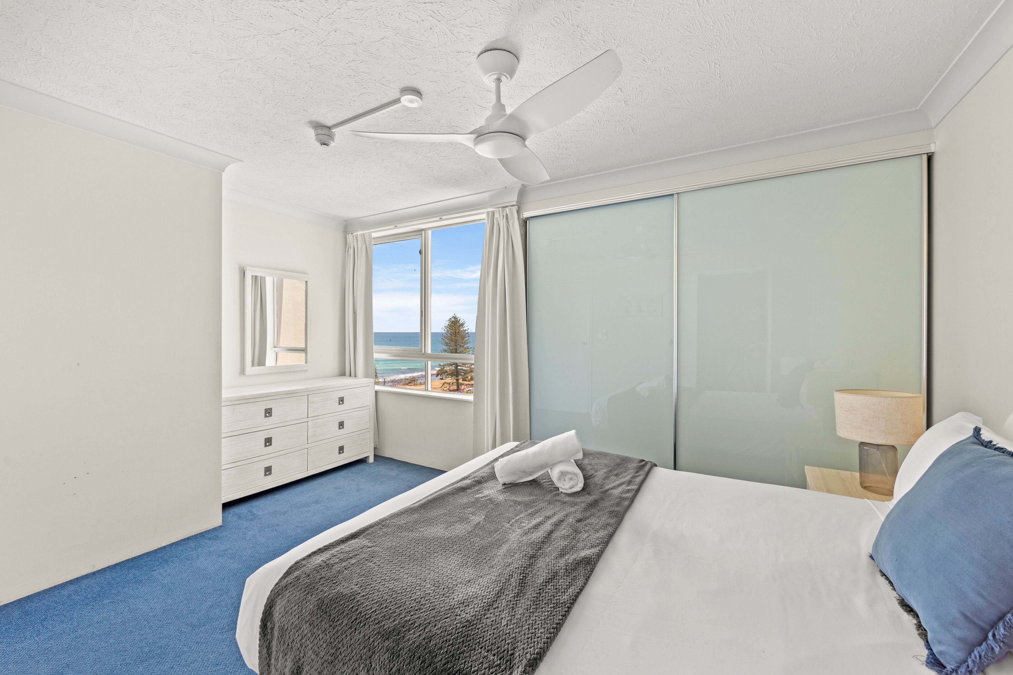 Une chambre à coucher comprenant un lit, un ventilateur de plafond, une commode, une fenêtre donnant sur la plage et une table de chevet avec une lampe.
