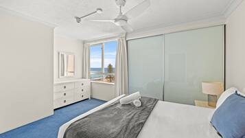 Une chambre à coucher comprenant un lit, un ventilateur de plafond, une commode, une fenêtre donnant sur la plage et une table de chevet avec une lampe.
