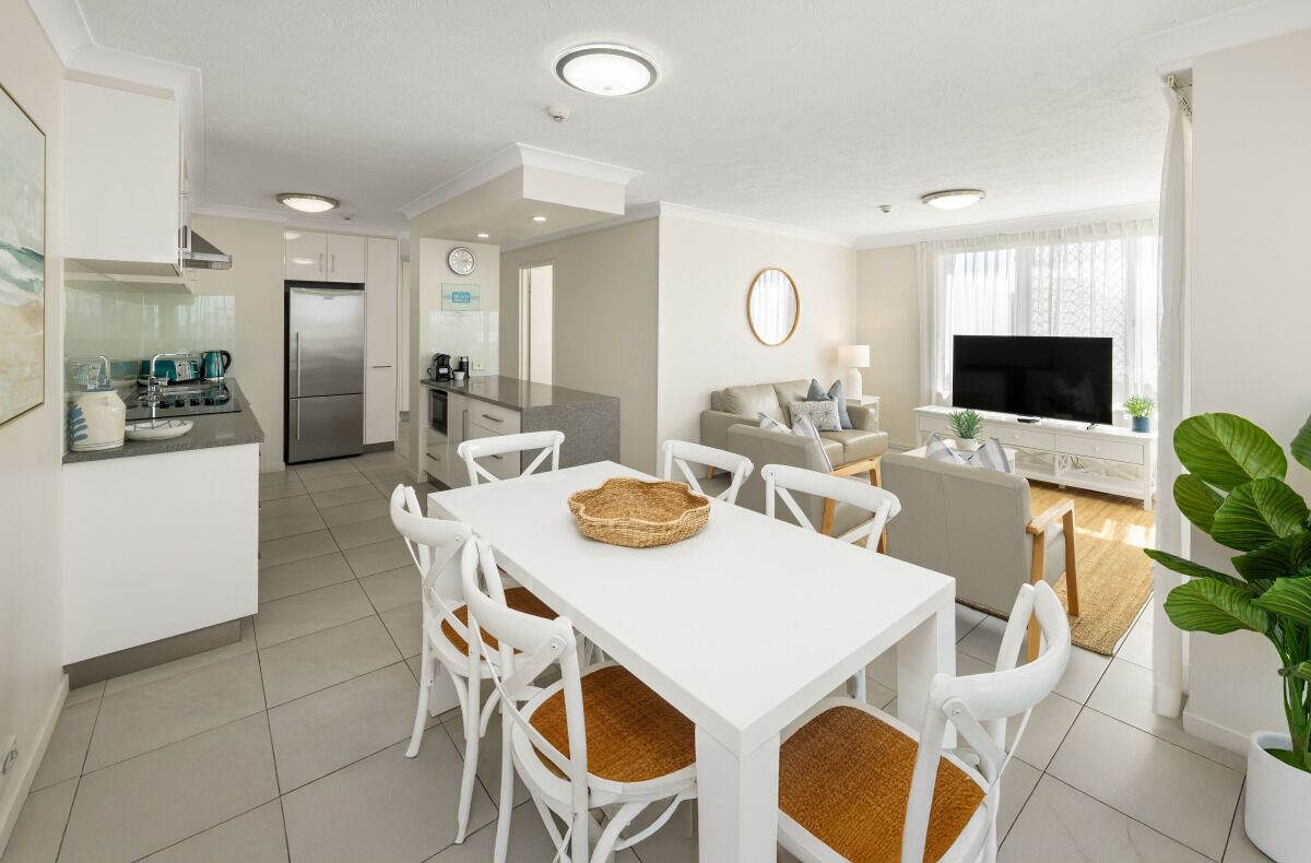 Een moderne keuken met witte kasten, een eettafel met stoelen, een televisie en een groot raam met gordijnen.