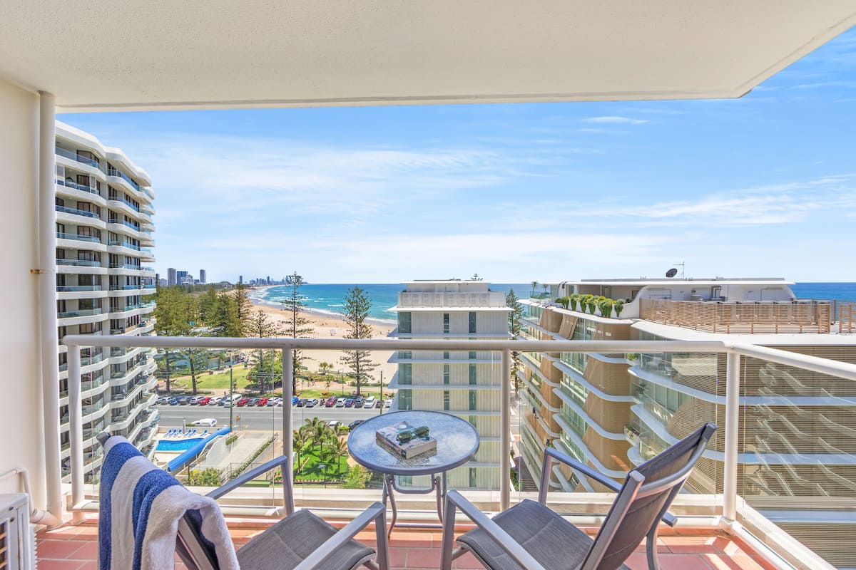A balcony with a view of the beach, a pool, and surrounding buildings.