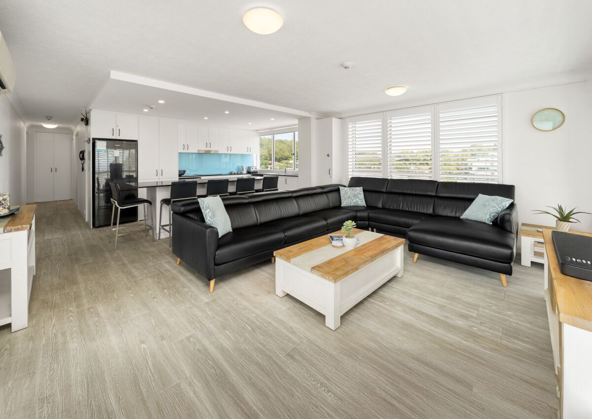 A modern living room with a black sectional sofa, a wooden coffee table, and a kitchen in the background.