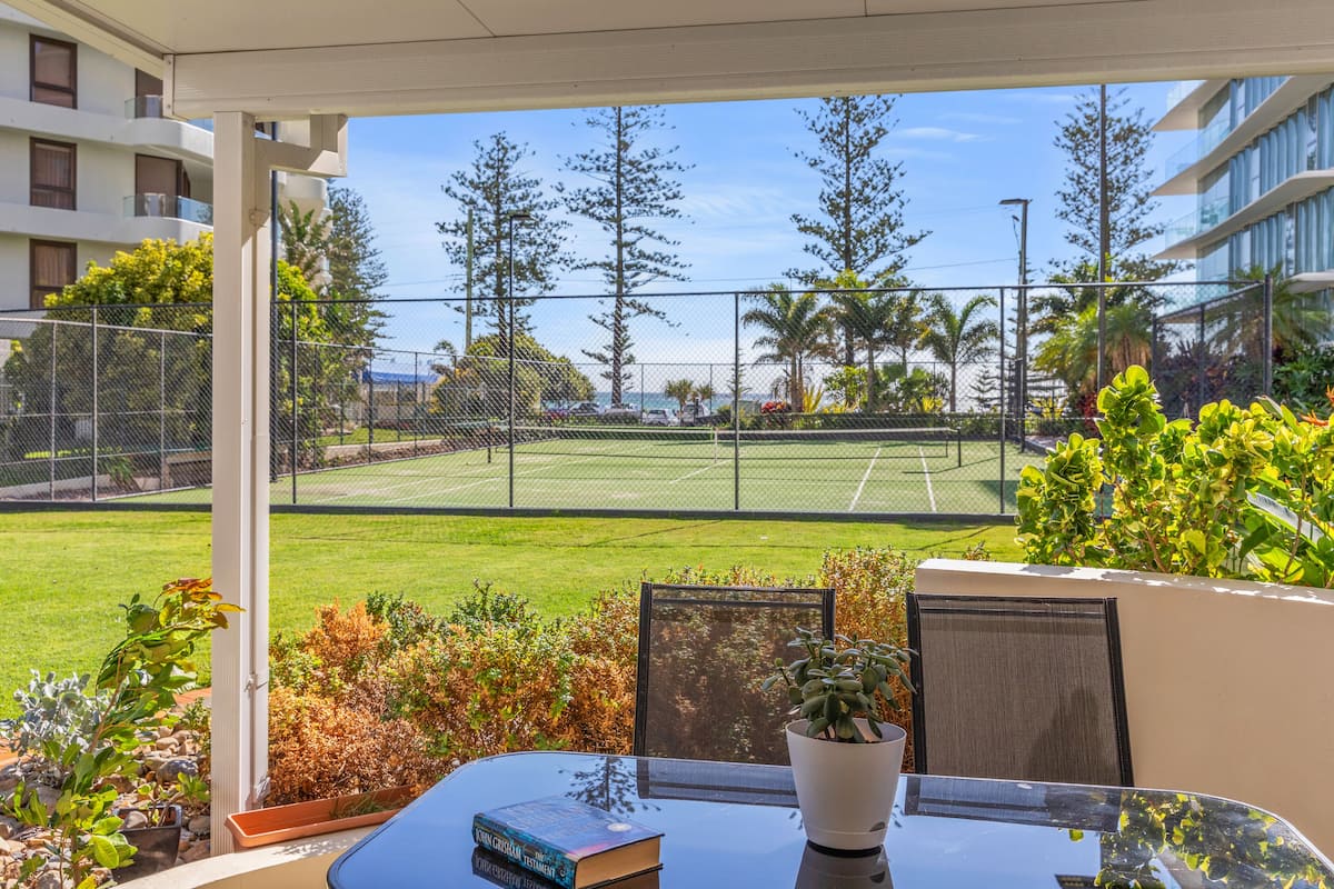 A balcony with a tennis court view, outdoor seating, and potted plants.