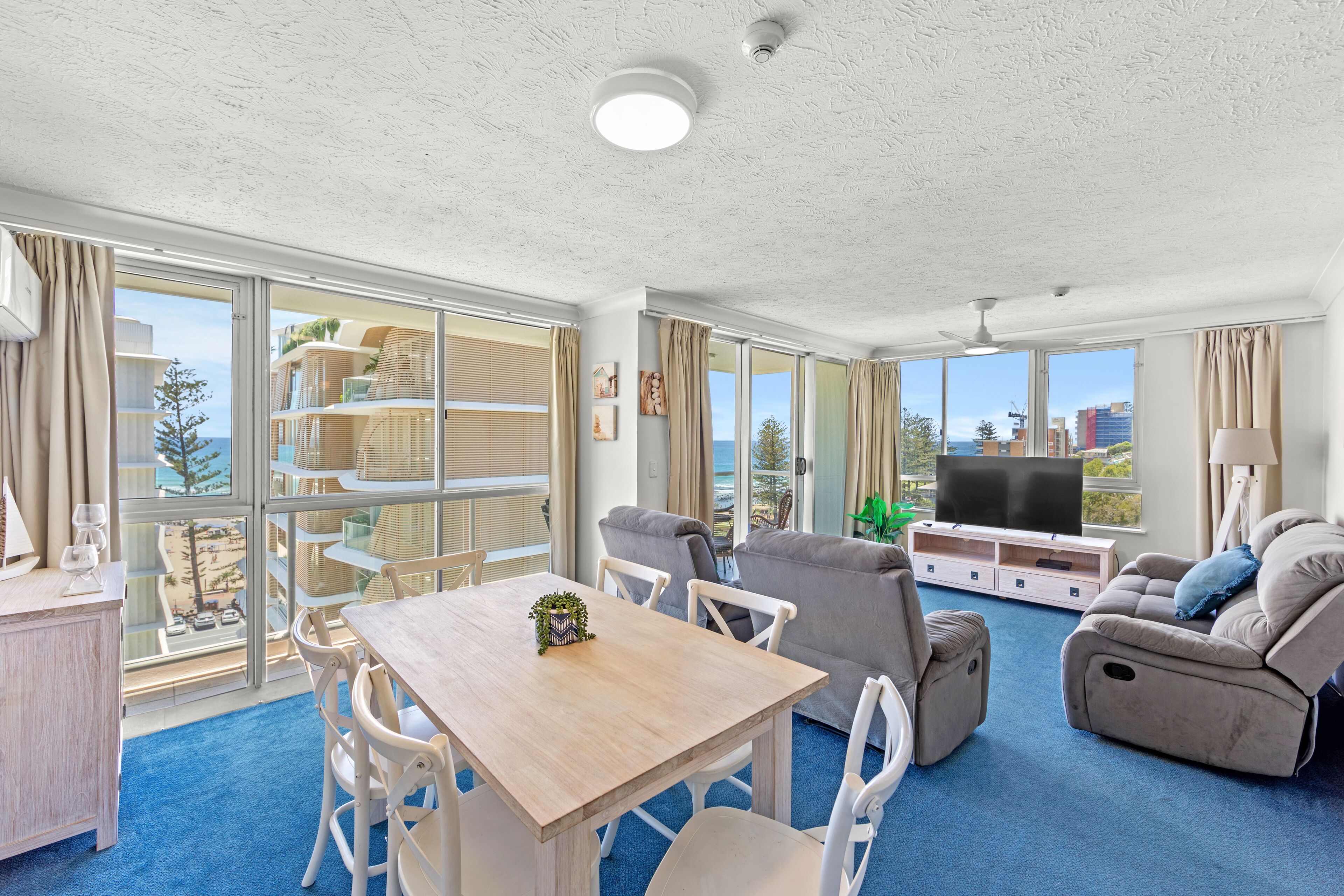 A spacious living area with a dining table, sofas, and a TV. Large windows offer a view of the city and the sea.