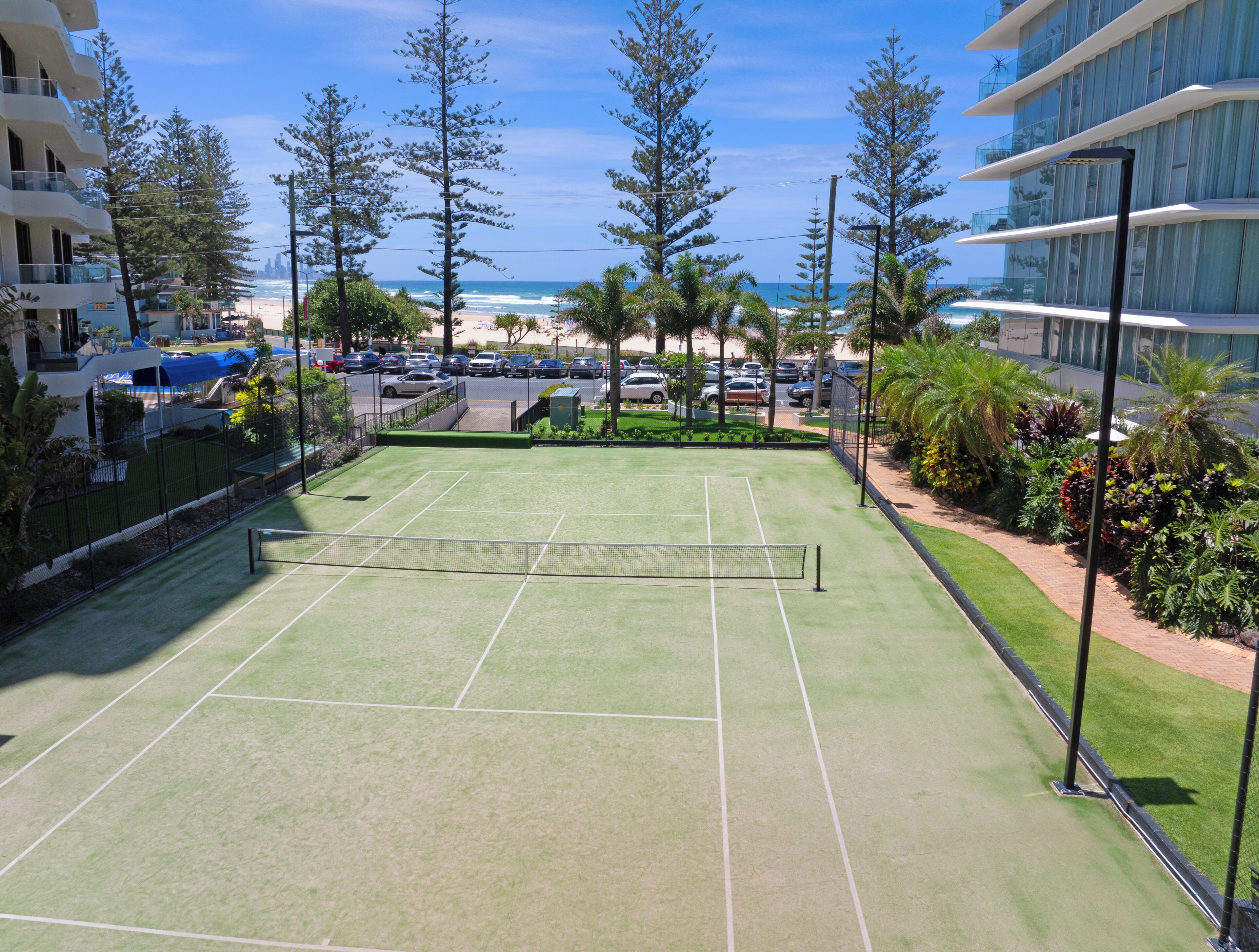 Ein Tennisplatz mit Netz, umzäunt, mit Blick auf einen Strand mit Palmen und einen Parkplatz.