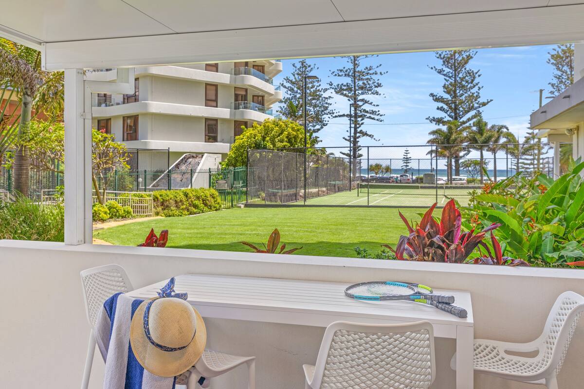 A balcony with a view of a tennis court, a white table, and a chair.
