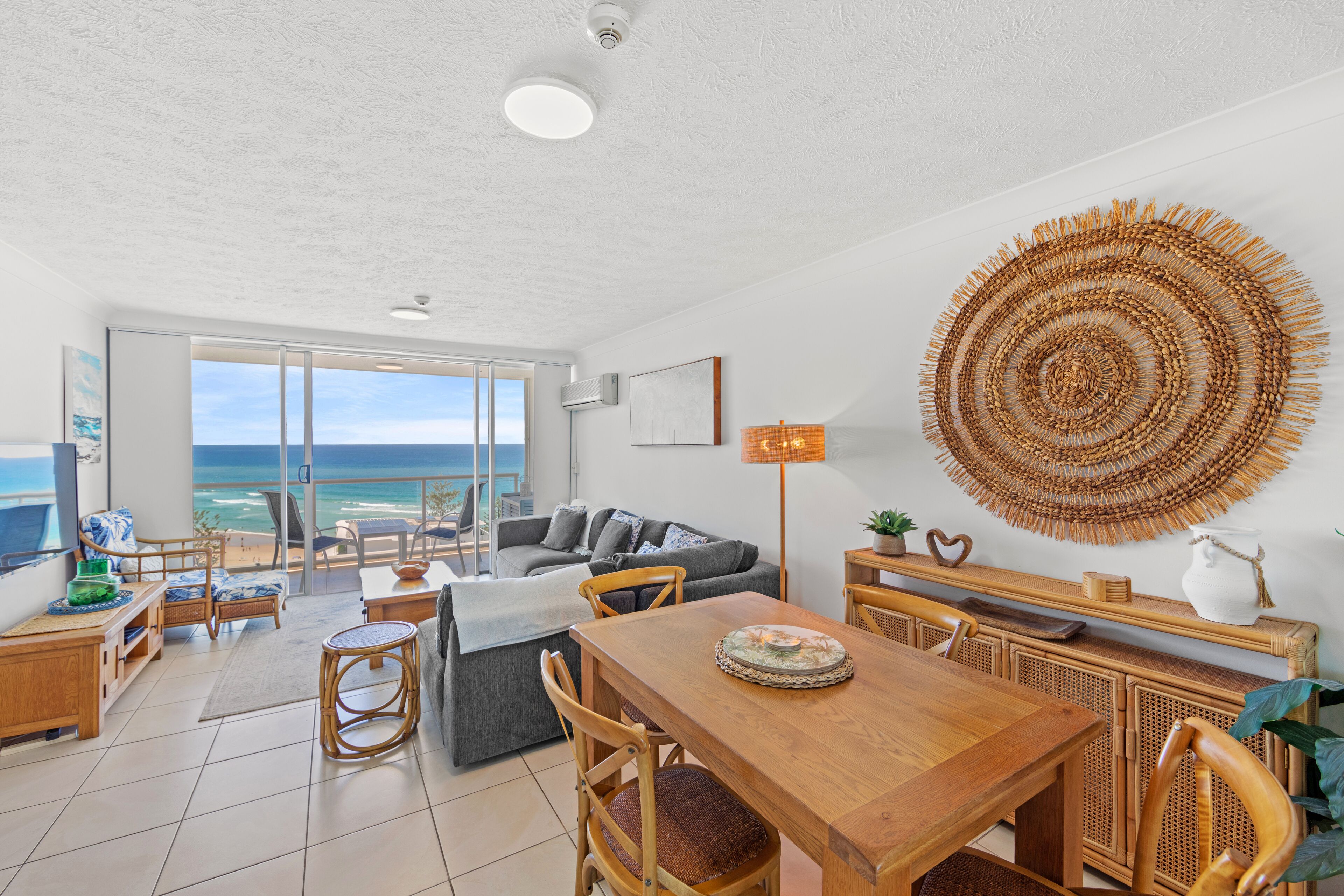 A spacious living area with a balcony overlooking the sea, a dining table, and a large woven wall hanging.