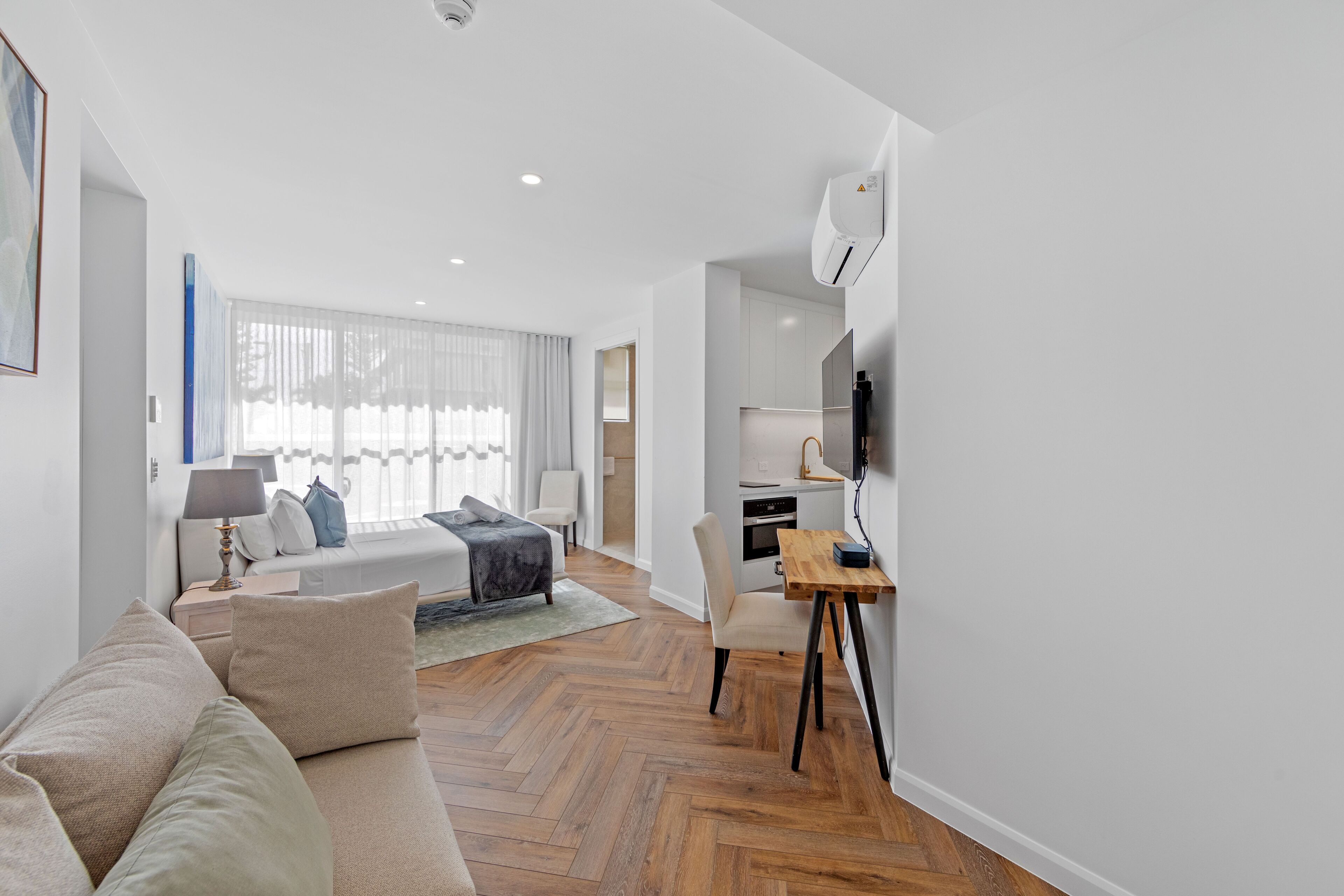 A modern hotel room with a wooden herringbone floor, a sofa, a dining table, and a TV mounted on the wall.