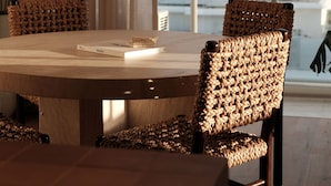 A dining area with a round table, wicker chairs, a hanging light fixture, and a view of the ocean through large windows. - Burleigh Beach Tower (Burleigh Heads)