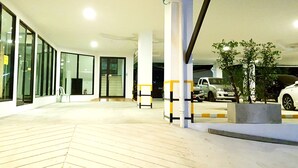 Entrada moderna de edifício com portas de vidro, piso de madeira e área de estacionamento com carros e uma árvore.