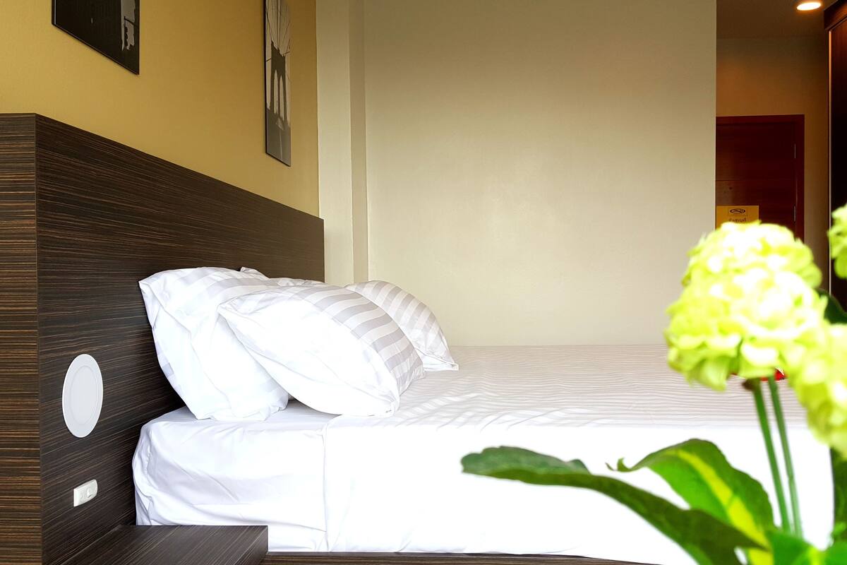 A neatly made bed with white bedding and pillows, a wooden headboard, a bedside table with a lamp, and a wall-mounted clock.
