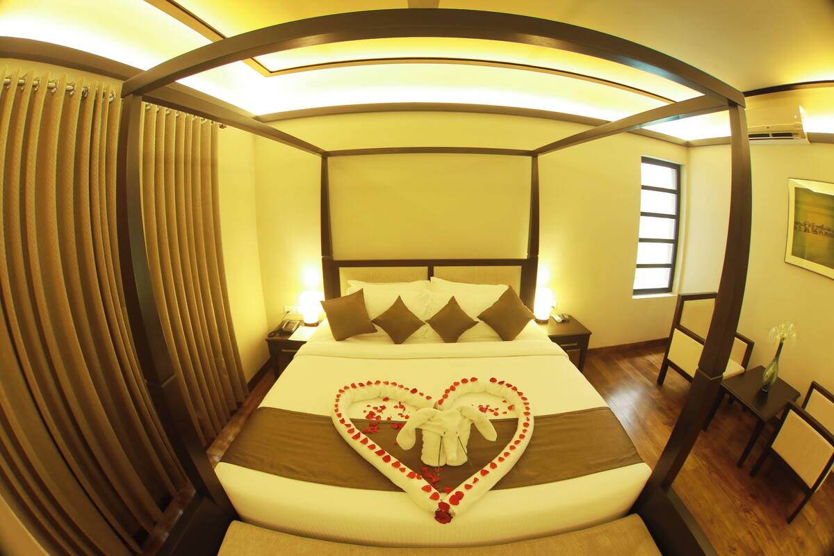 A hotel room with a large bed, a heart-shaped decoration on the bed, a nightstand, a chair, and a window.