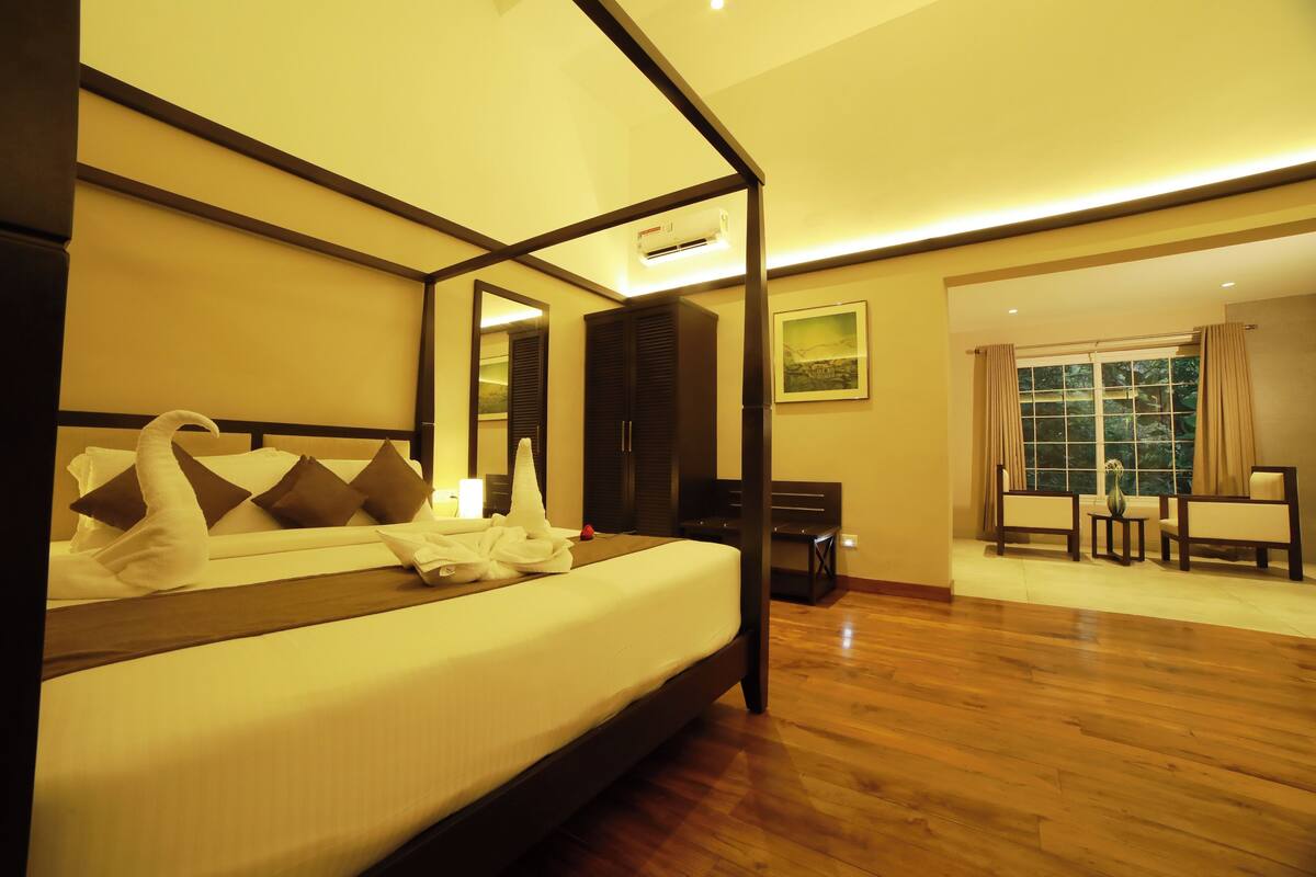 A hotel room with a four-poster bed, a sitting area with a table and chairs, and a large window with curtains.