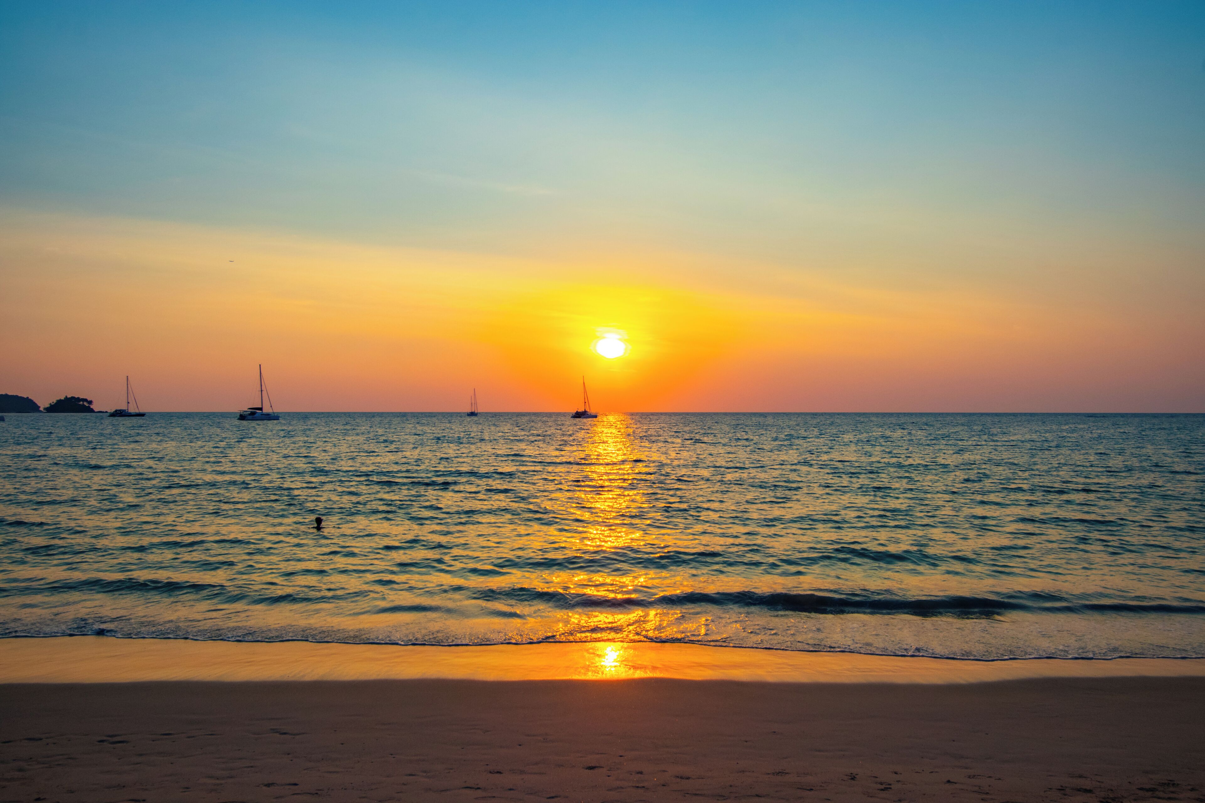 A sunset over a calm sea with boats, a sandy beach, and a clear sky.