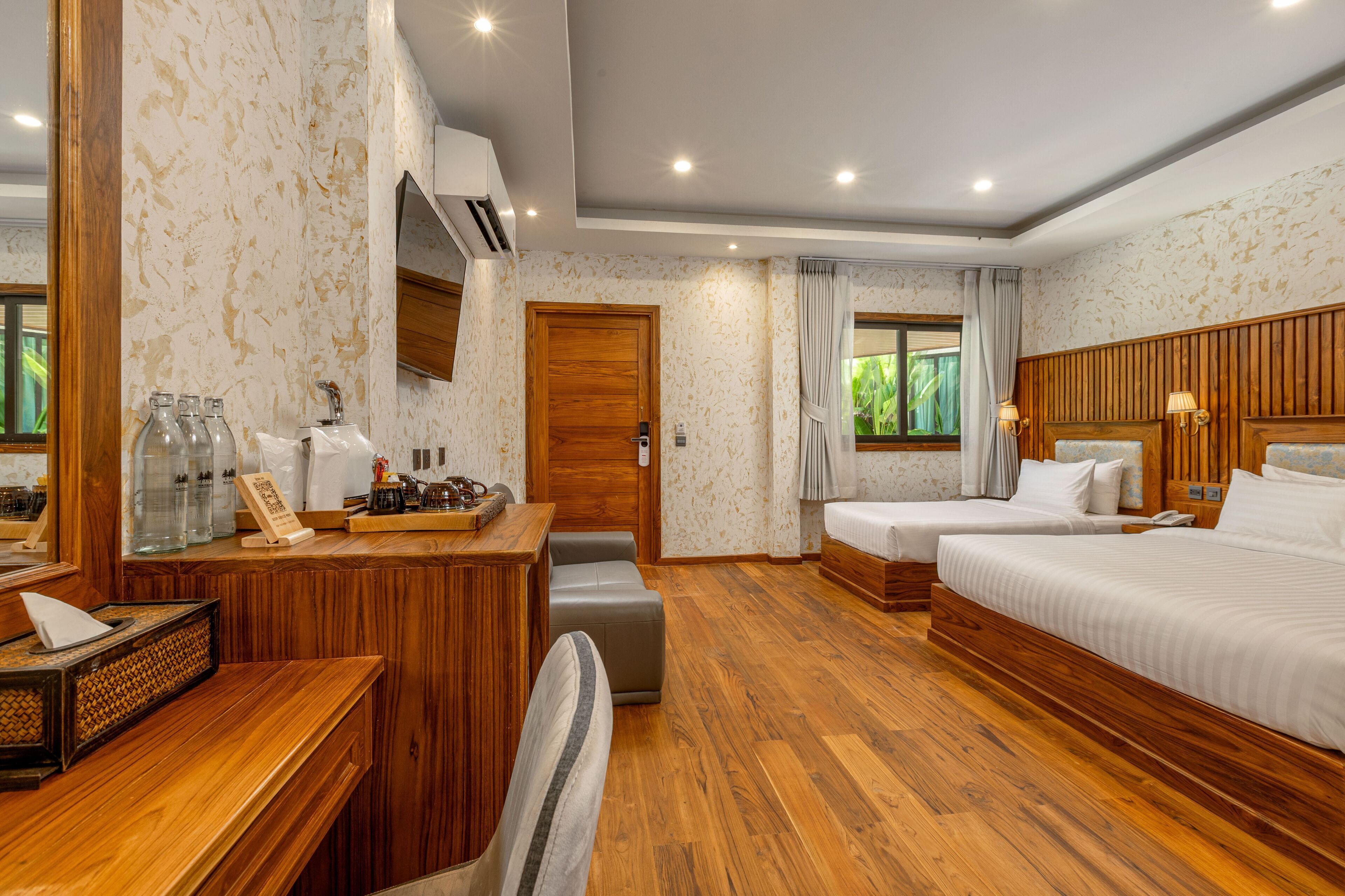 A hotel room with a wooden desk, two beds, a window with curtains, and a wooden headboard.