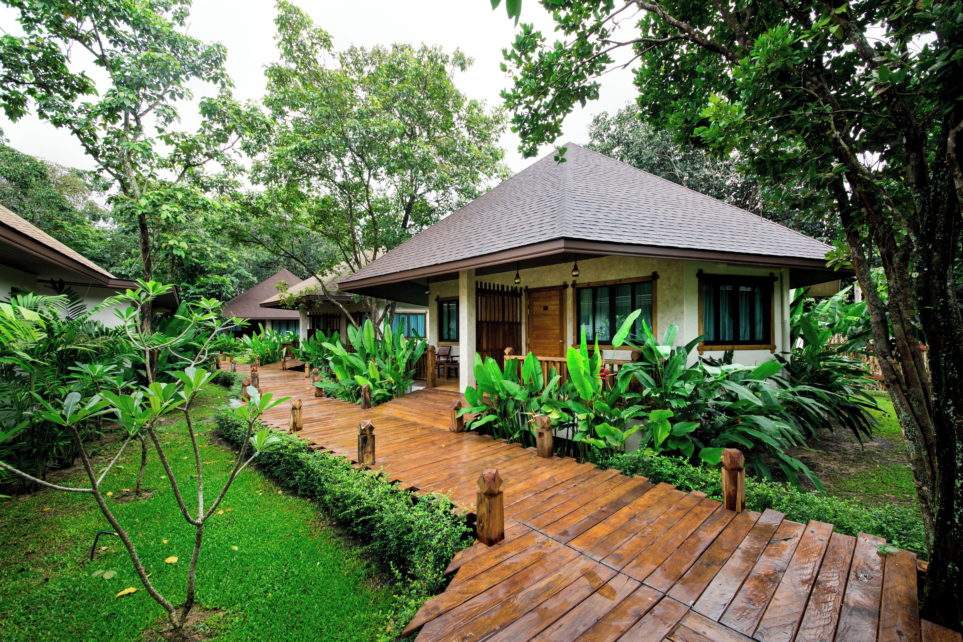Un sentier en bois mène à un bungalow doté d’un toit en tuiles, entouré d’une végétation luxuriante et de plantes tropicales.