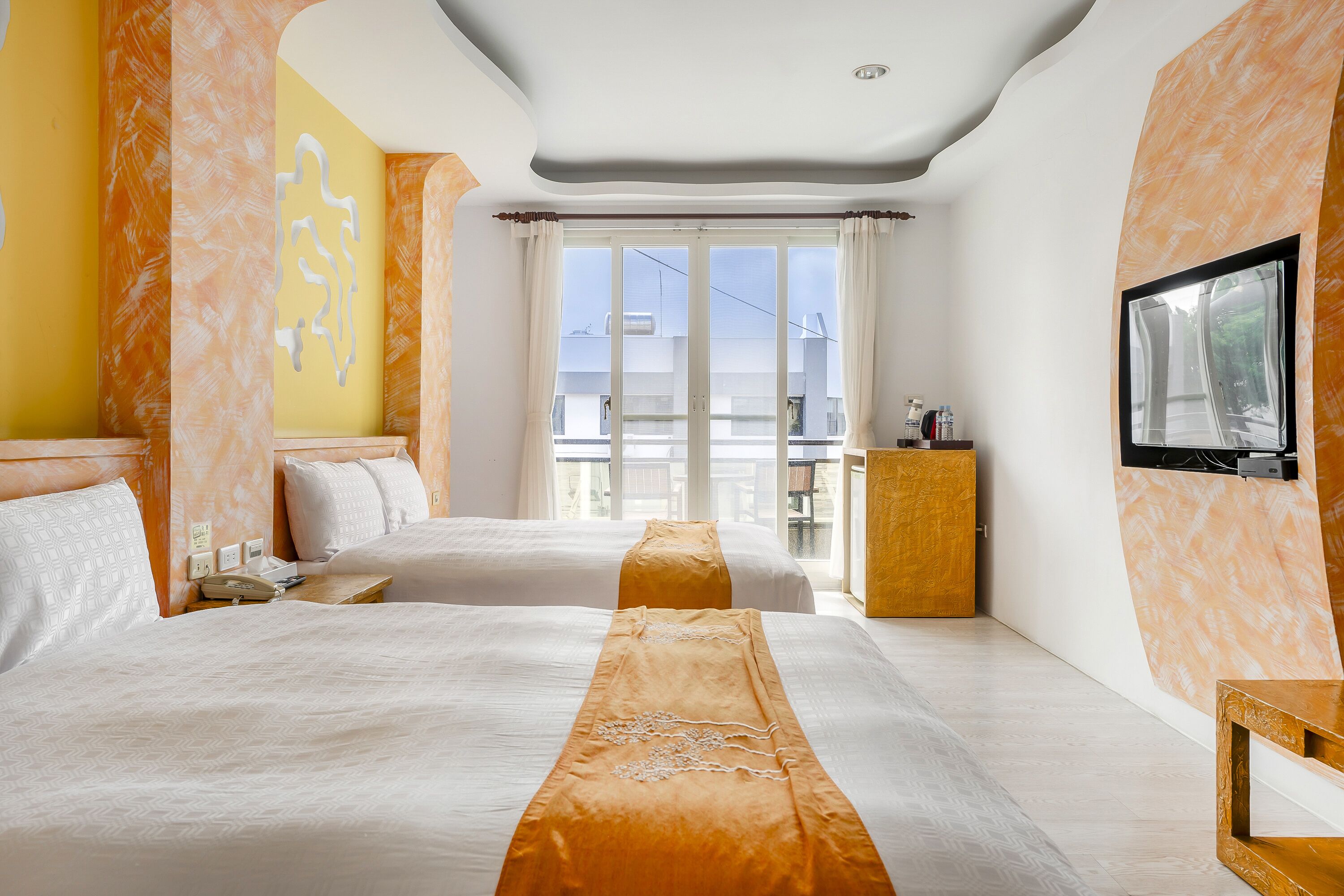 A hotel room with two beds, a TV mounted on the wall, and a balcony with a view.