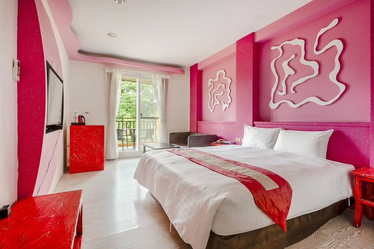 A modern hotel room with a large bed, a red chair, a desk, and a balcony with a view of greenery.