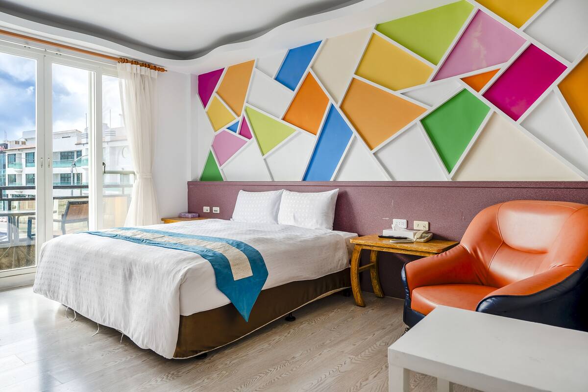 A hotel room with a bed, a chair, a small table, and a wall with colorful geometric patterns.