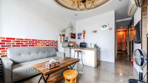 A modern living room with a grey sofa, wooden coffee table, and a wicker stool. A kitchen area with a bar stool and a fan is visible in the background. - Wei Yuan Hotel (Hengchun)