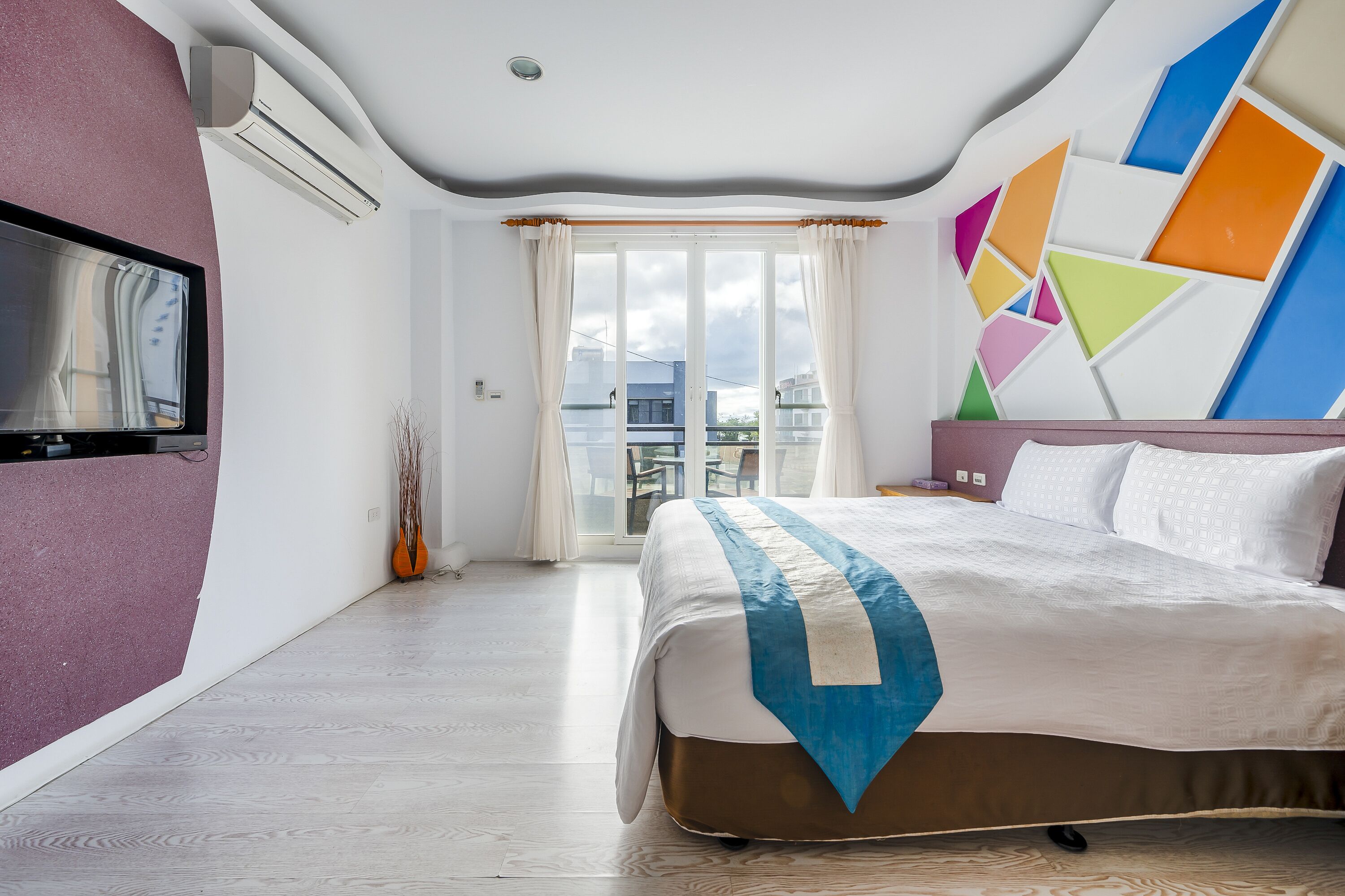 Habitación de hotel con una cama grande, un televisor, un balcón con vistas y un diseño geométrico en la pared.