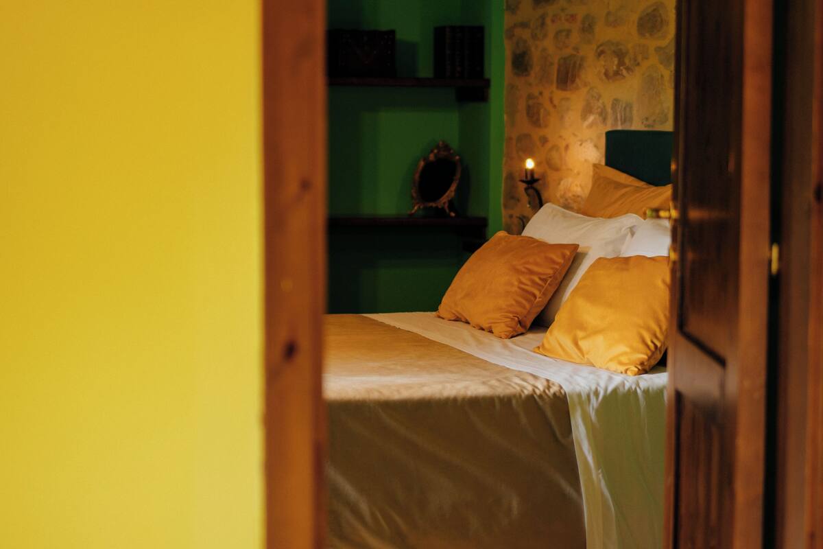 A bed with white and yellow bedding, a bedside table with a mirror and a candle, and a green wall with shelves in the background.